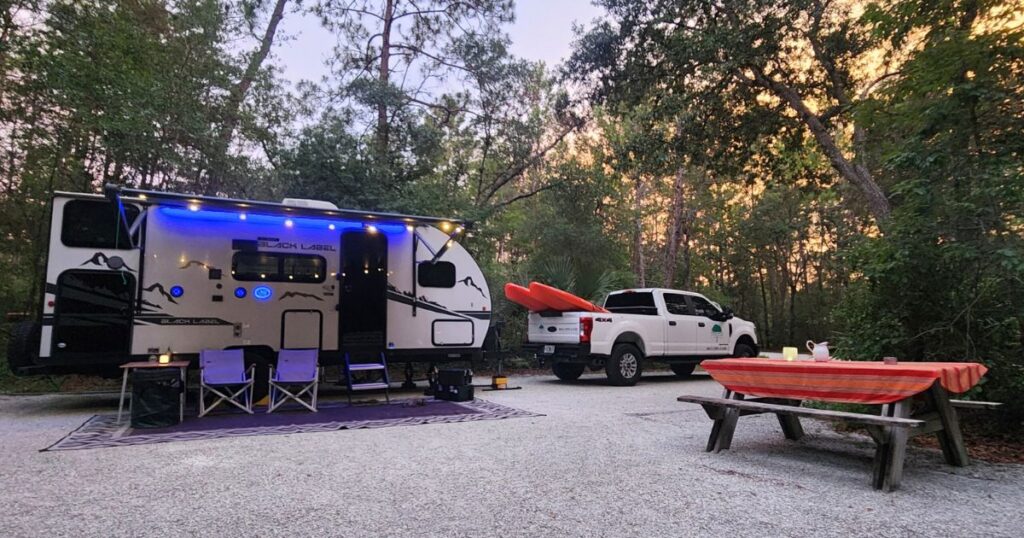 Cozy campsite with a travel trailer, outdoor seating, and kayaks in nature. Perfect for adventure and outdoor family fun.