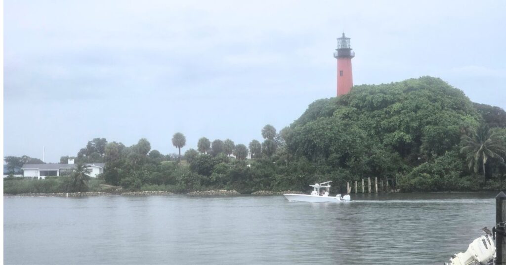 Lighthouse on lush green island near water with boat and cloudy sky.