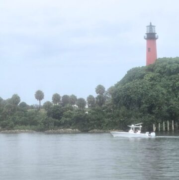 Lighthouse on lush green island near water with boat and cloudy sky.