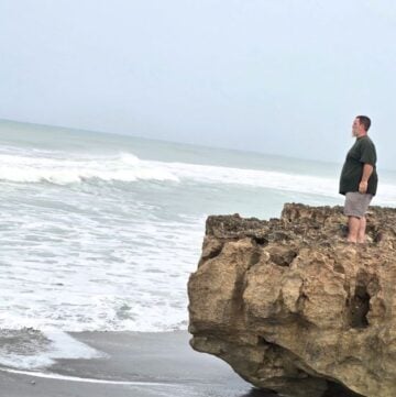 Seaside landscape with man standing on rocky shoreline, ocean waves crashing, overcast sky, Hawaii coast, contemplative moment.
